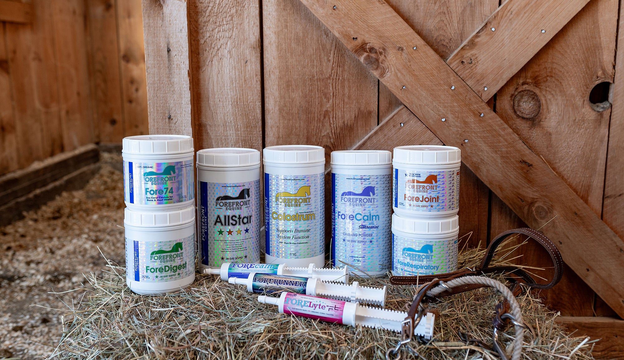 The comprehensive ForeFront Equine Supplements collection displayed in a rustic stable setting features specialized formulas including ForeDigest, ForeJoint, AllStar, Colostrum, and ForeCalm, all scientifically formulated to enhance equine health, performance, and recovery in competitive and recreational horses.